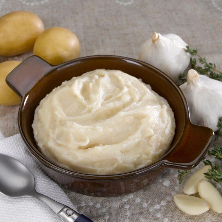 Delicious protein mashed potatoes served in a bowl, surrounded by garlic, thyme, and fresh potatoes.